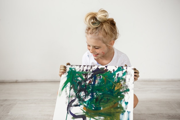 Free photo joyful, cheerful, smiling with her teeth and looking down at her picture little blonde. european female child wearing white t-shirt sitting on the floor and holding picture.