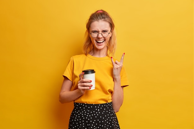 Joyful confident relaxed girl makes heavy metal sign, enjoys rock, laughs happily, holds coffee to go, spends free time on wild concert, awesome music festival