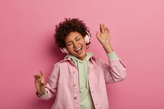 Joyful curly haired teenage woman dances carefree listens audio track in headphones