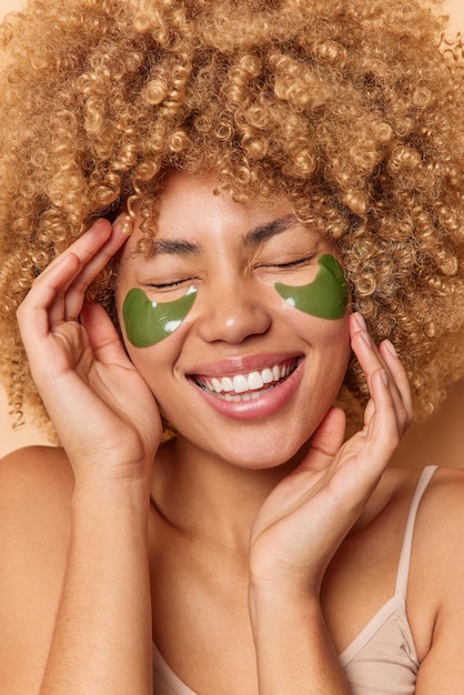 Joyful curly haired woman laughs out happily keeps eyes closed wears hydrogel green patches from dark circles smiles toothily enjoys beauty treatments touches face expresses positive emotions.