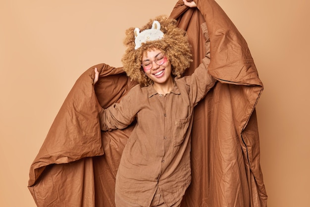 Free photo joyful curly haired young woman dressed in pajama sleepmask on forehead holds soft blanket has fun before sleeping applies beauty patches isolated over brown background. good morning concept