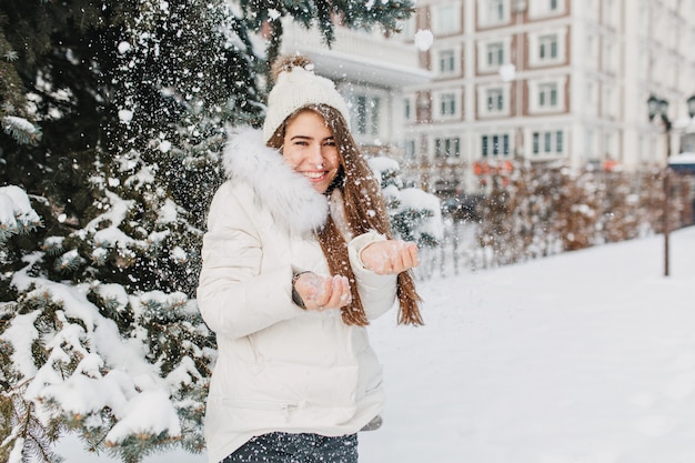 Free photo joyful cute woman having fun with snowflakes outdoor on fir tree full with snow. young charming model in warm winter clothes enjoying cold snowing on street. expressing positivity, smiling.