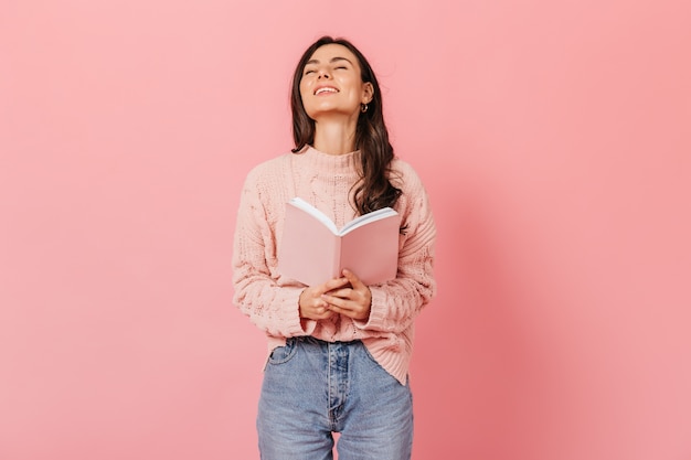 Free photo joyful dark-haired woman laughs while reading book. portrait of dark-haired girl in light sweater on pink background.