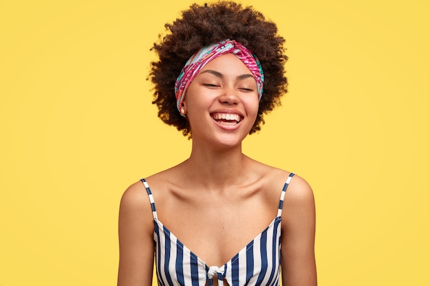 Free photo joyful dark skinned model laughs pleasantly, closes eyes from happiness, recieves wonderful suggestion, being in high spirit during summer trip, wears headband and striped top, poses indoor.