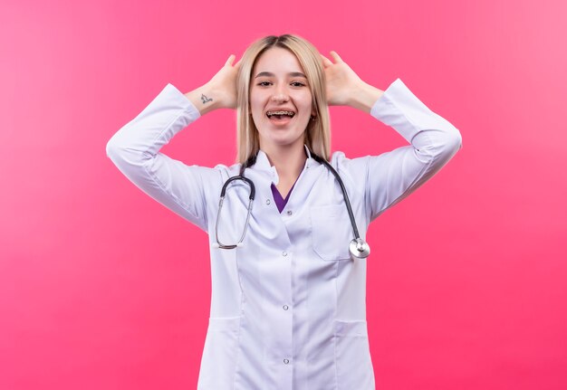 Joyful doctor young blonde girl wearing stethoscope in medical gown and dental brace put her hands on head on isolated pink wall