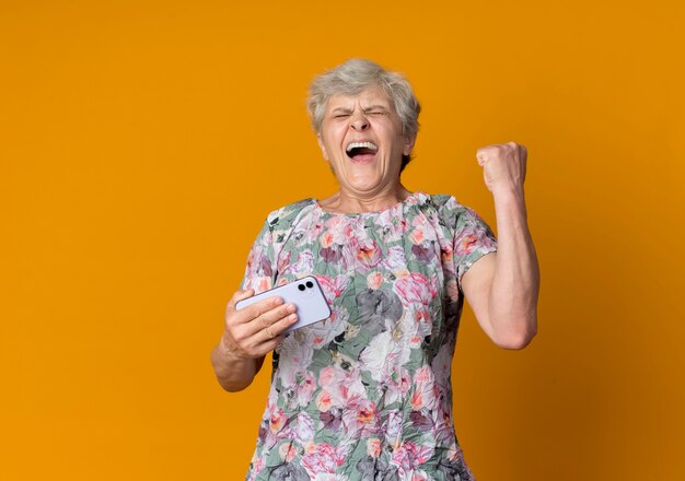 Joyful elderly woman holds phone and raises fist up isolated on orange wall