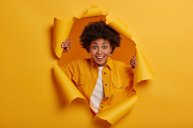 Free Photo joyful ethnic woman feels happy, stands through torn hole of yellow background