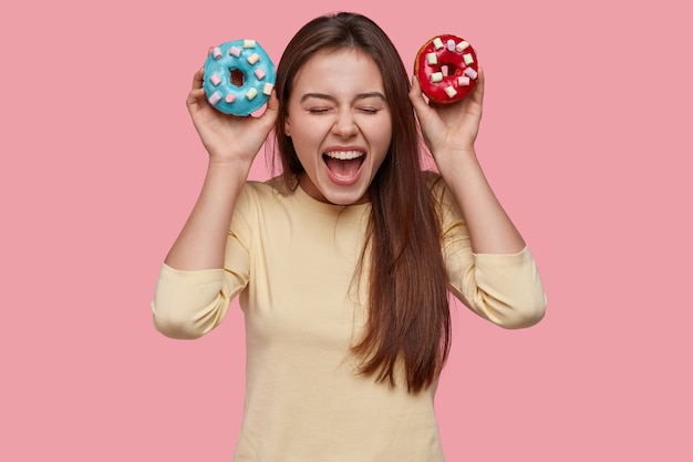 Joyful excited lady exclaims from happiness, carries two blue and red donuts, has tempatation to eat it, keeps to diet, opens mouth broadly