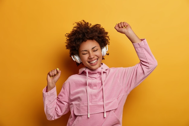 Free photo joyful expressive female listens music in headphones, enjoys pleasant melodies, has good mood, dances carefree, smiles broadly, wears pink sweatshirt, poses against yellow wall. people, leisure