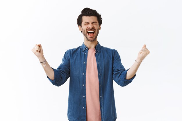 Joyful extremely happy handsome bearded guy yelling from happiness out loud, close eyes and clench fists, pump hands up from joy, winning feeling lucky, triumphing, celebrating amazing victory