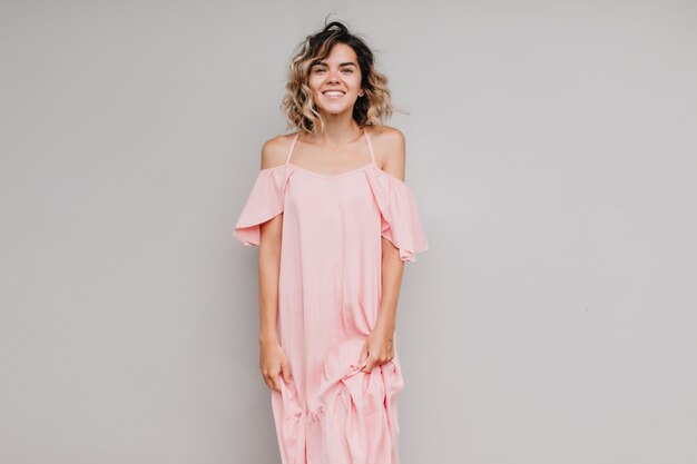 Joyful female model with curly hair posing with sincere smile. Indoor photo of tanned girl in long pink dress isolated.