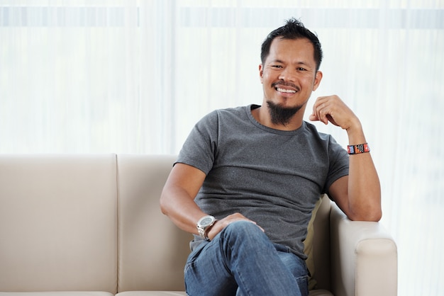 Joyful Filipino man seated on couch smiling contentedly at camera