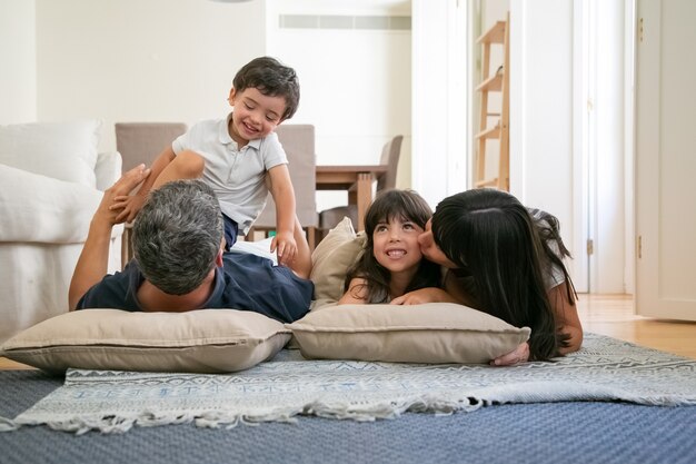 Joyful funny parents cuddling, hugging and kissing little kids, having fun together