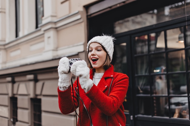 Free photo joyful girl in red jacket, knitted hat and mittens takes picture of city with retro camera.