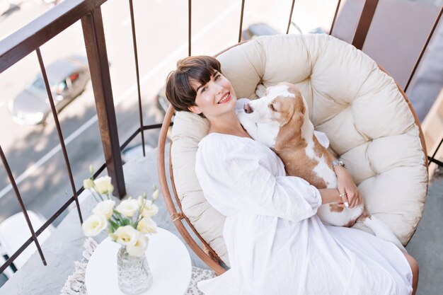 Joyful girl sitting in chair beside vase of white roses and stroking beagle dog. Beautiful brown-haired woman enjoying fresh air on balcony with pet lies on her knees