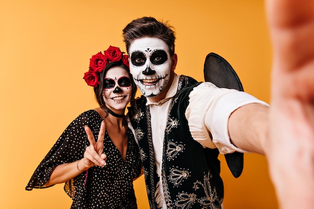 Free photo joyful guy and girl are enjoying halloween party. couple takes selfie in unusual clothes showing peace sign