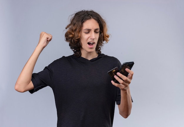 Joyful guy with long hair in black t-shirt holding a phone and a card on white wall