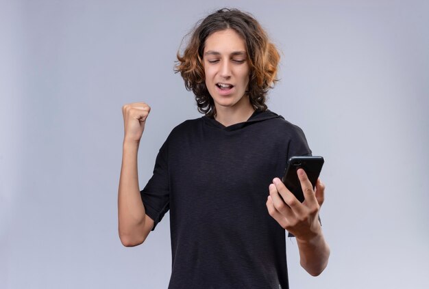 Joyful guy with long hair in black t-shirt holding a phone on white wall