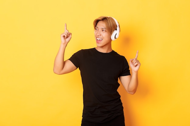 Joyful handsome asian guy with blond hair, singing along and dancing as listening to music in wireless headphones, standing yellow wall