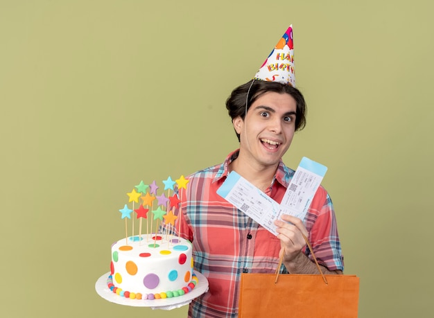 Joyful handsome caucasian man wearing birthday cap holds paper shopping bag air tickets and birthday cake 