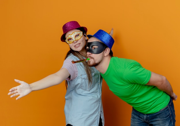 Free photo joyful handsome man in blue hat wearing masquerade eye mask blowing whistle looking at side an surprised young girl wearing pink hat and masquerade eye mask pens arms looking