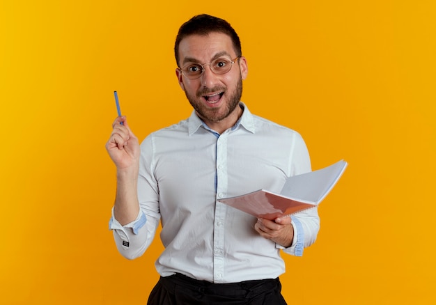 Joyful handsome man with optical glasses holds pen and notebook isolated on orange wall