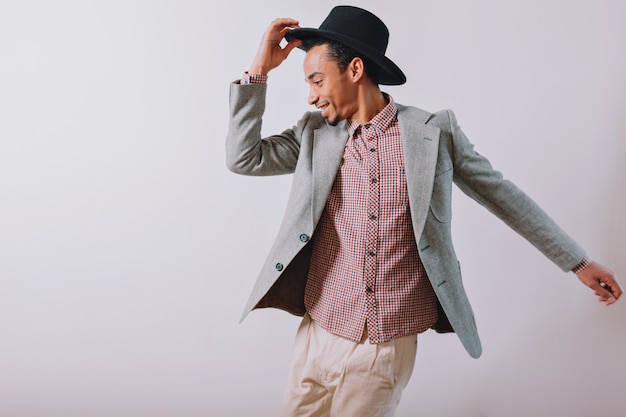 Free photo joyful happy man wears black hat and grey suit dancing with excited emotions on gray