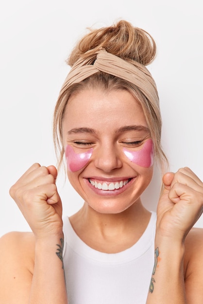 Joyful happy woman clenches fists rejoices good news undergoes beauty treatments applies hydrogel patches enjoys effective beauty treatment wears beige headband isolated over white studio wall