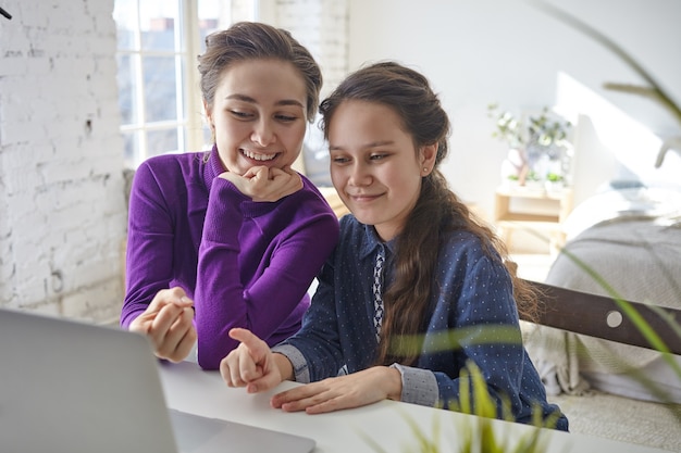 Joyful happy young mother and daughter shopping online using laptop pc, sitting at desk in light bedroom interior, pointing fingers at screen and smiling