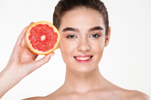 Joyful image of smiling half-naked woman with natural makeup holding orange citrus near her face and looking