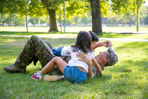 Joyful kids and their dad lying and playing on grass in park. Happy military father meeting with children after mission trip. Family reunion or returning home concept