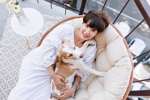 Joyful lady in bathrobe with short hairstyle posing in chair with dog sitting near vase with flowers.