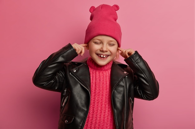 Free Photo joyful little preschooler covers ears, keeps index fingers in ear holes, avoids hearing loud music, has happy expression, wears pink hat with ears and leather jacket, doesnt want to hear noise