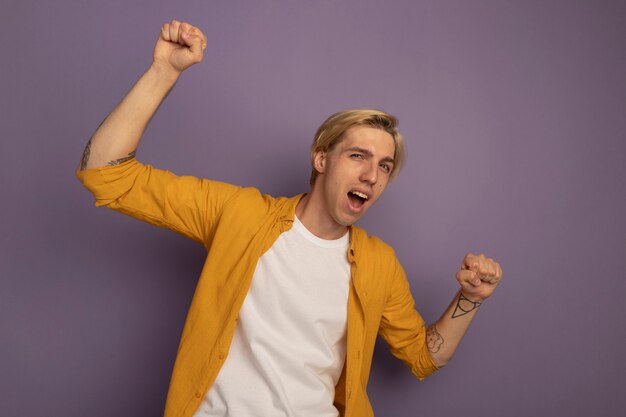 Joyful looking straight ahead young blonde guy wearing yellow t-shirt showing yes gesture isolated on purple