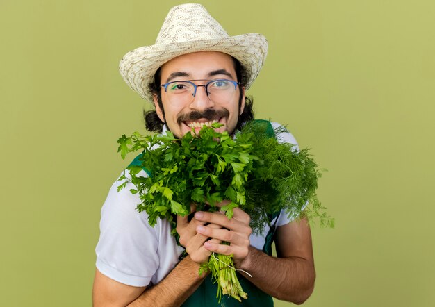 Joyful male gardener in optical glasses wearing gardening hat holds fennel and coriander 