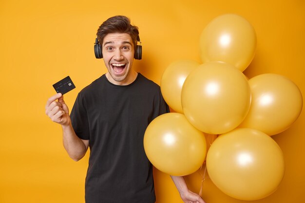 Joyful man happy to get big sum of money on his bank account ready to celebrate birthday holds bunch of inflated balloons and credit card listens music via headphones isolated over yellow wall