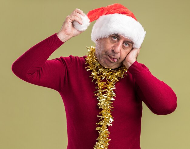 Joyful middle age man wearing christmas santa hat with tinsel around neck looking at camera touching his hat sticking out tongue standing over green  background