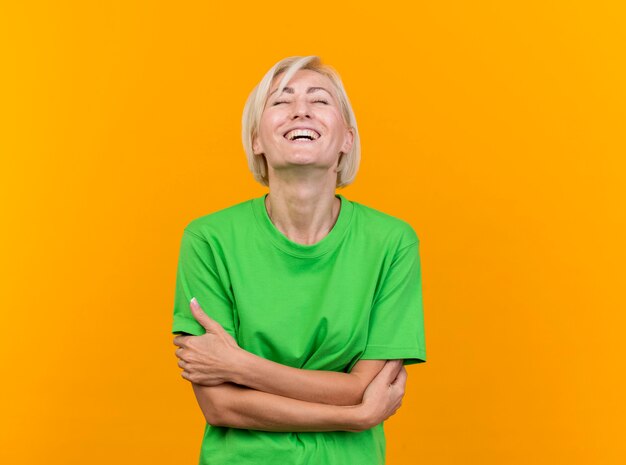 Joyful middle-aged blonde slavic woman standing with closed posture and laughing with closed eyes isolated on yellow background with copy space