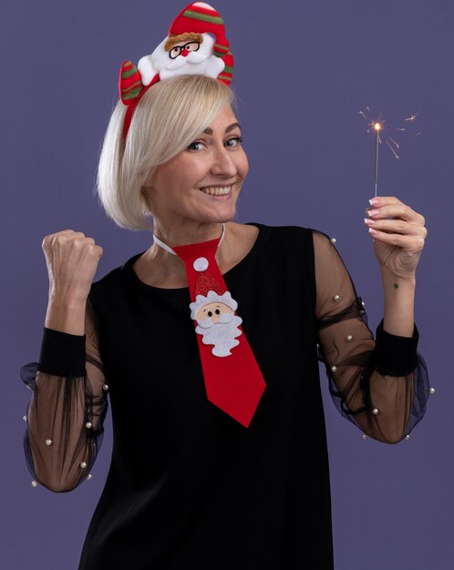 Joyful middle-aged blonde woman wearing santa claus headband and tie holding holiday sparkler  doing yes gesture isolated on purple wall