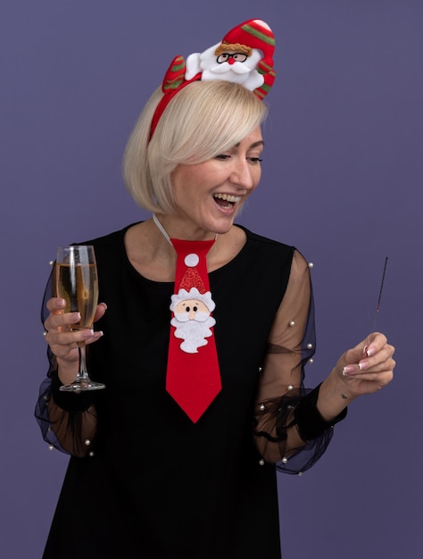 Joyful middle-aged blonde woman wearing santa claus headband and tie holding holiday sparkler and glass of champagne looking at sparkler isolated on purple wall