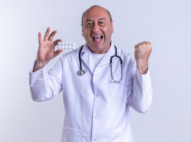 joyful middle-aged male doctor wearing medical robe and stethoscope showing pack of tablets looking at front doing yes gesture isolated on white wall