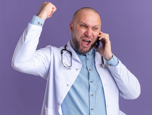 Free Photo joyful middle-aged male doctor wearing medical robe and stethoscope talking on phone looking at side doing yes gesture isolated on purple wall