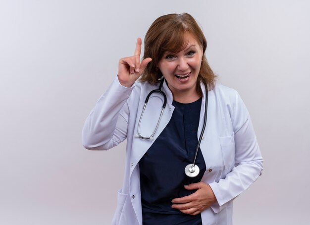 Joyful middle-aged woman doctor wearing medical robe and stethoscope pointing with finger up and putting hand on belly on isolated white wall with copy space