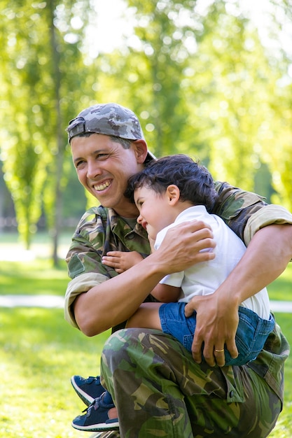 Joyful military dad holding little son in arms, hugging boy outdoors after returning from mission trip. Vertical shot. Family reunion or returning home concept