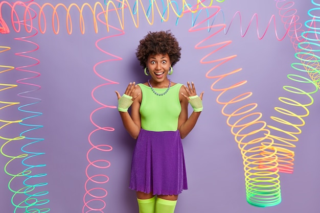 Free Photo joyful millennial girl raises hands, wears sport gloves, fashionable colorful outfit, fools around, has fun on party, poses indoor with colorful slinky toys around. youth