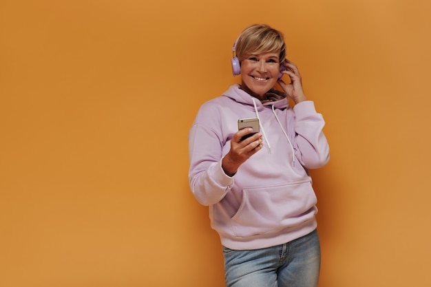 Free photo joyful old woman with stylish blonde hairstyle and lilac headphones in trendy pink hoodie and jeans smiling and holding smartphones.