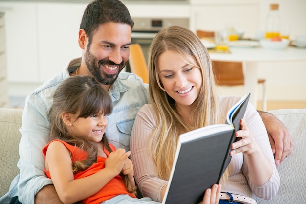 Joyful parents couple and little black haired girl sitting on couch in living room, reading book together and laughing.