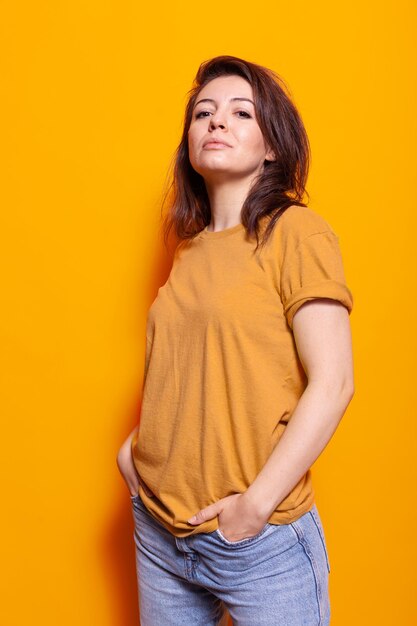 Joyful person standing over orange background in studio. Cheerful woman posing with confidence, feeling casual while looking at camera. Stylish adult smiling and having hands in pockets