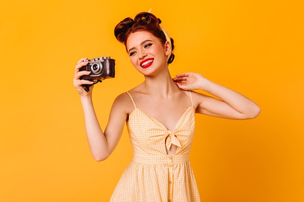 Joyful pinup girl in dress taking photos. Studio shot of elegant laughing woman with camera isolated on yellow space.