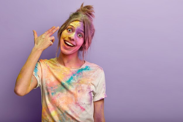 Joyful pleased young female shoots in temple, demonstrates finger gun gesture, suicide sign, tilts head, has colorful t shirt and face smeared with powder on Holi holiday, blank space aside for text